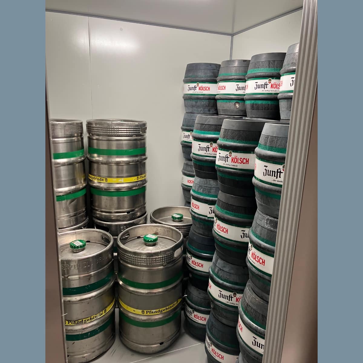 Metallfässer stapeln sich in einem Kühlraum, viele Fässer mit grünem Band und Kölsch-Etiketten, rechter Fassstapel hoch, linke Bereiche mit runden Metallbehältern