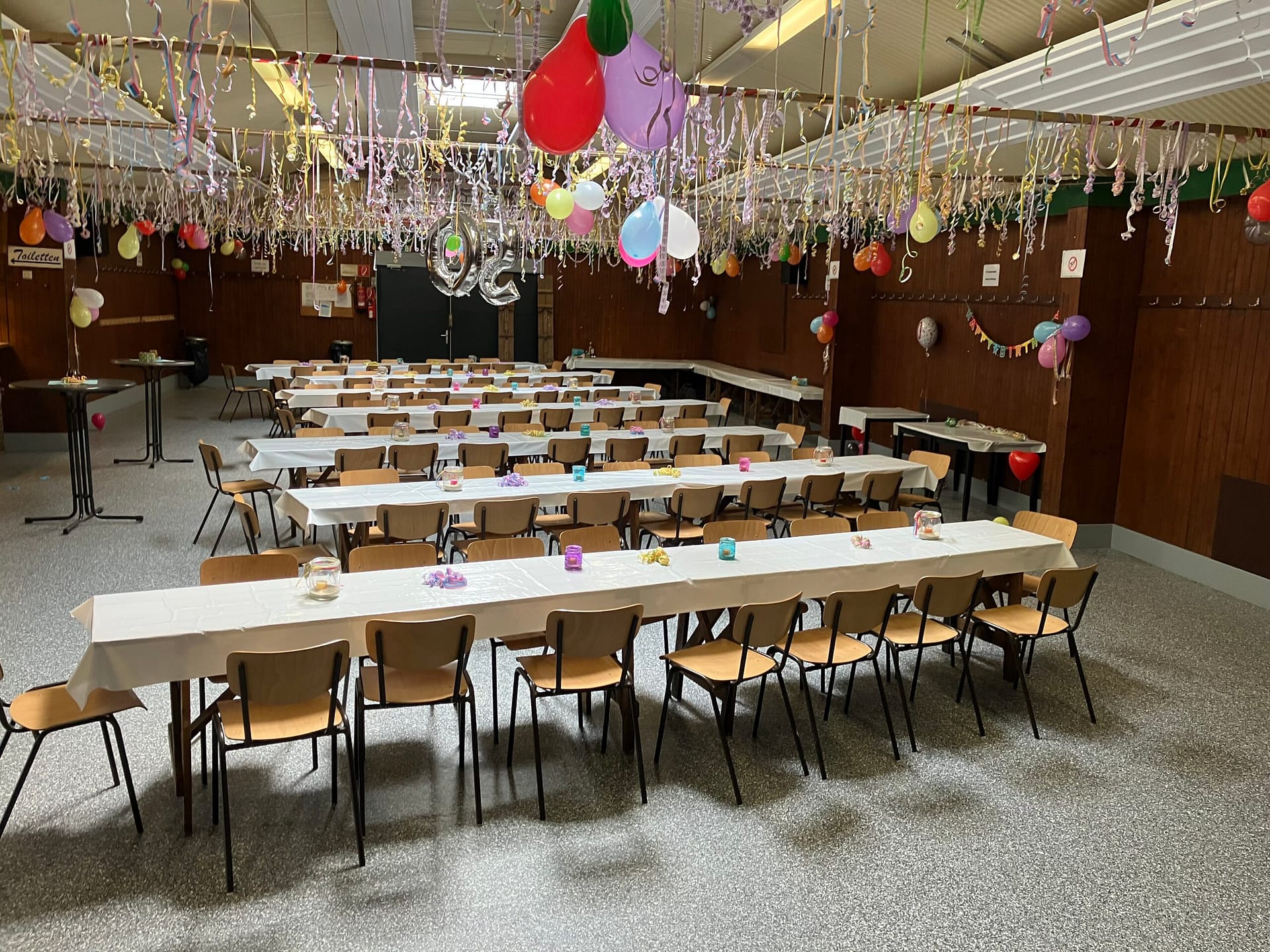 Großer Veranstaltungsraum mit langen Tischen und bunten Luftballons sowie Dekorationen für eine Feier.