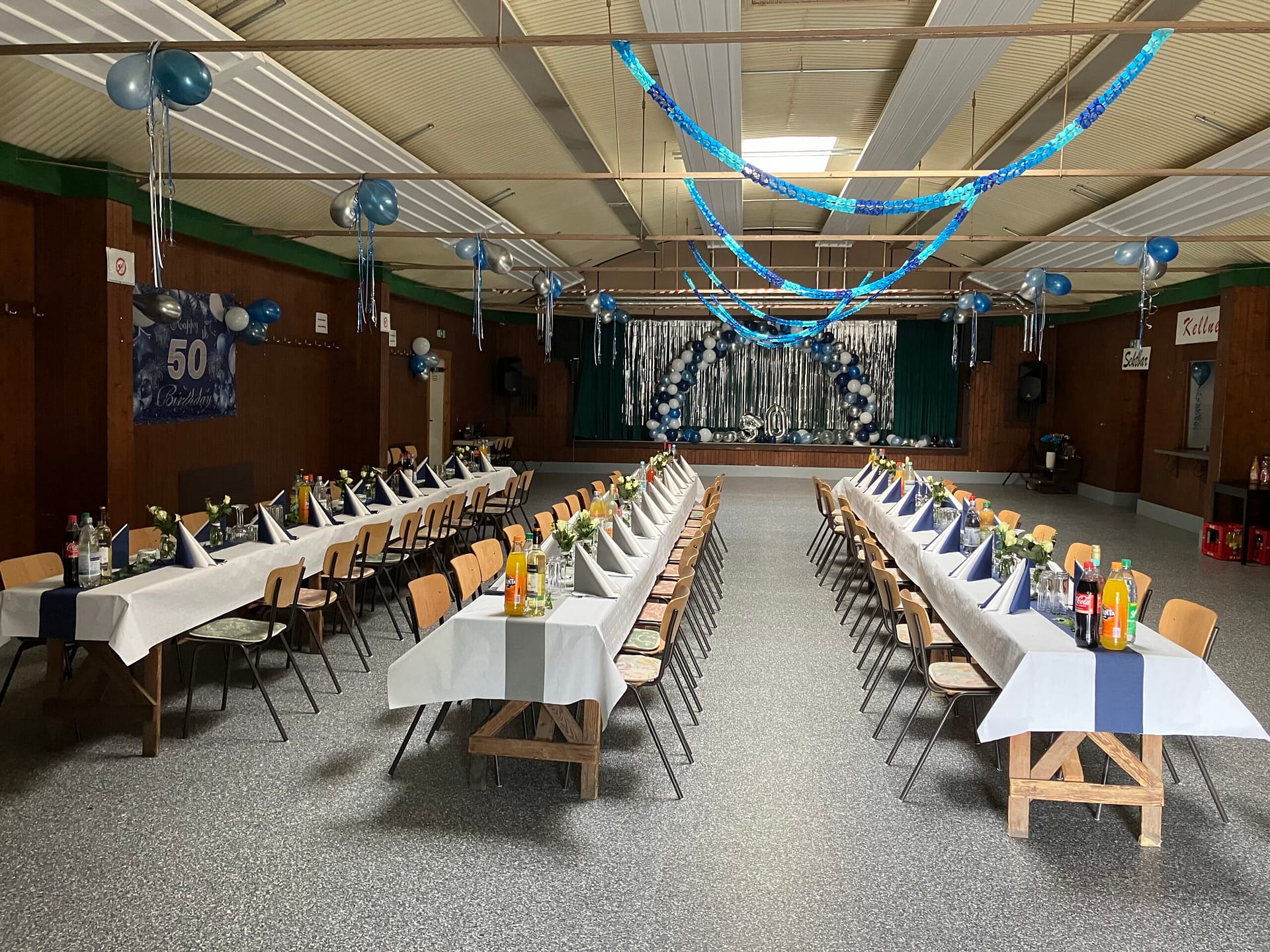 Festlich dekorierter Raum mit Tischen, Stühlen und blauen Luftballons für eine Feier zum 50. Jubiläum.