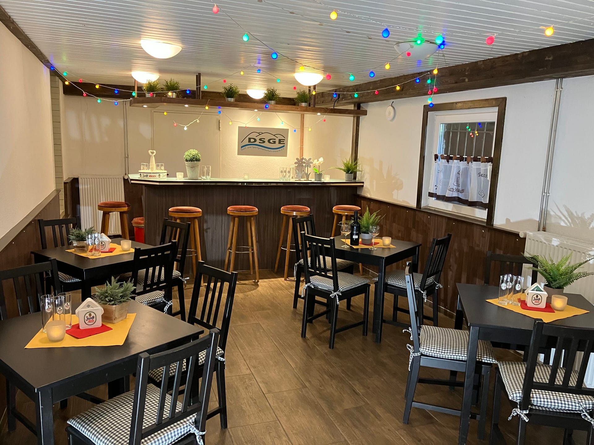 Gemütlicher Innenraum eines Restaurants mit Tischen, Stühlen und einer Bar im Hintergrund, dekoriert mit bunten Lichtern.