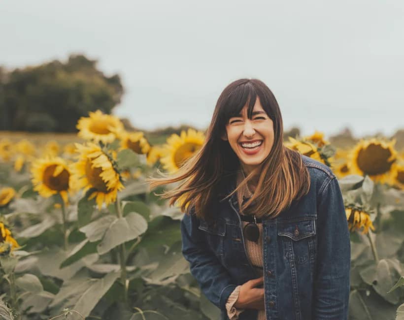 In the Present for You campaign, a laughing woman stands in a sunflower field on a cloudy day, wearing a denim jacket and sweater.