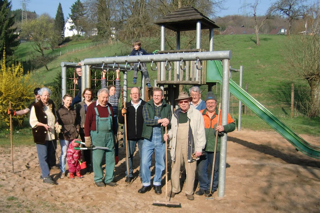 Eine Gruppe von Personen steht vor einem Spielplatz mit einem Spielgerät und Werkzeugen in der Hand.