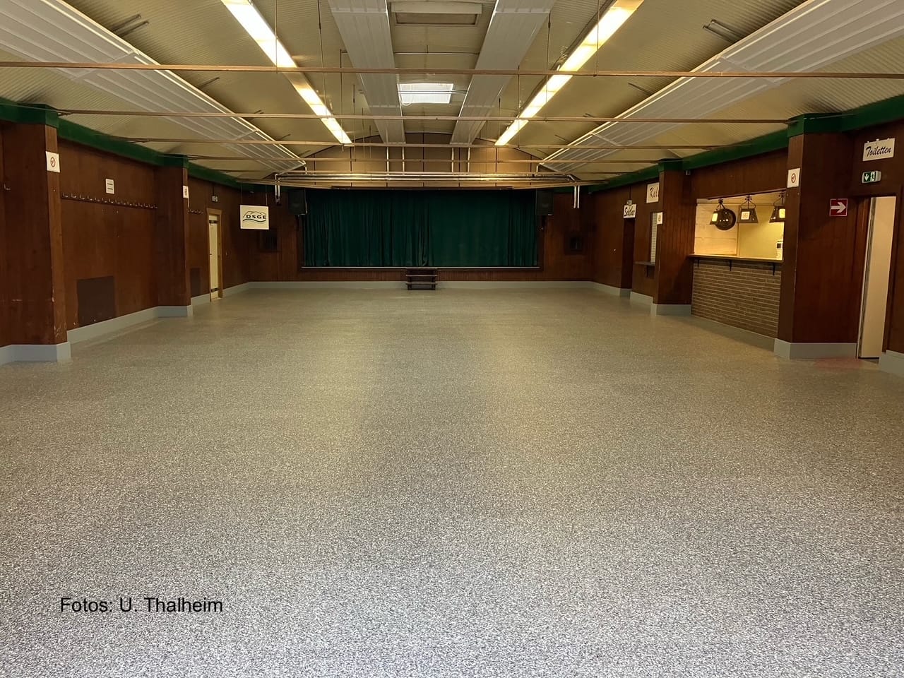 Innenansicht eines großen, leeren Veranstaltungsraums mit grauem Bodenbelag und Holzverkleidung.