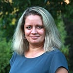 Frau mit blonden Haaren und grauem Oberteil, die in die Kamera lächelt.