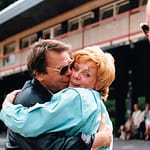 Ein Mann umarmt eine Frau mit blonden Haaren und einer türkisfarbenen Jacke, beide lächeln in einer freundlichen Umarmung.