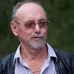 Mann mit Brille und Bart, trägt ein gestreiftes Hemd und eine schwarze Weste, lächelt leicht in die Kamera.