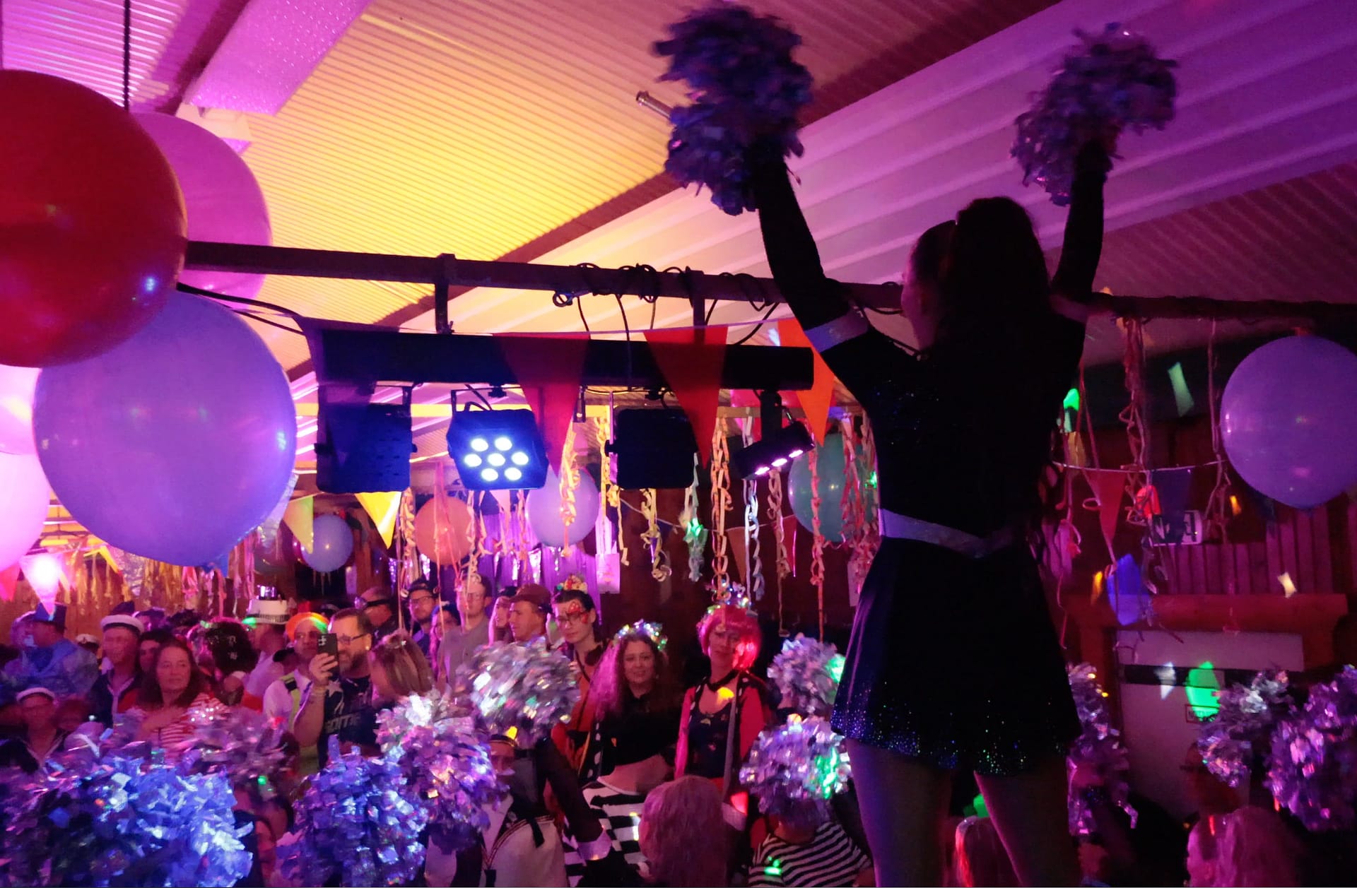 Eine Person mit Pompons steht auf einer Bühne während einer Feier mit bunten Lichtern und Dekorationen.