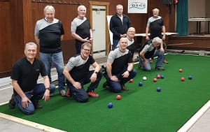 Gruppe von Männern in Sportkleidung, die auf einem Boule-Platz stehen und Boule-Kugeln werfen.