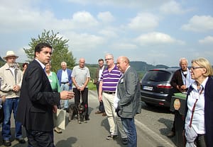 Mehrere Dorfbewohner stehen bei einer Begehung auf einer Landstraße, während ein Mann im Anzug spricht.