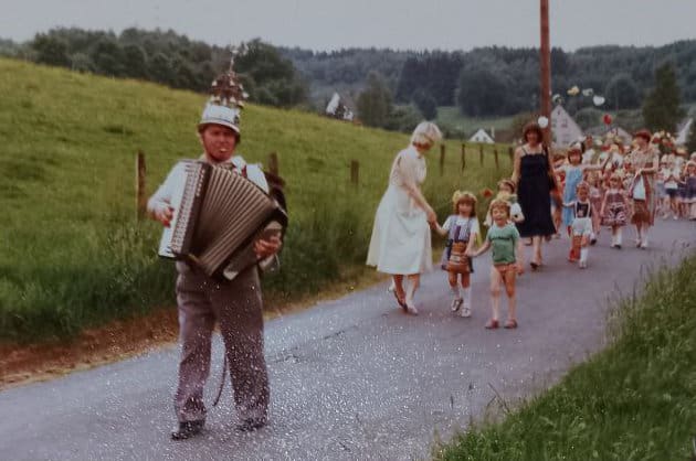 Mann mit Akkordeon in traditioneller Kleidung, der eine Gruppe von Menschen bei einem Umzug anführt.